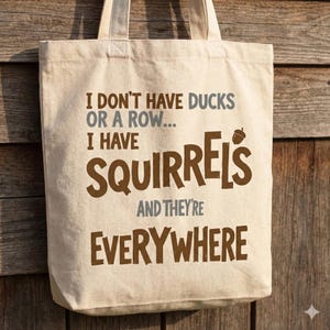 May include: A beige canvas tote bag with the text "I don't have ducks or a row... I have squirrels and they're everywhere" in brown and gray lettering. The bag has two handles and is hanging against a wooden background.