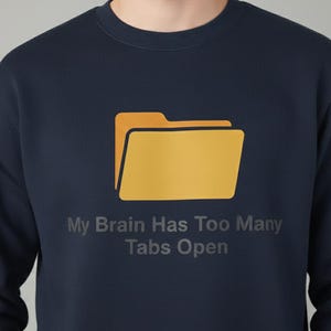 Op de afbeelding: Marineblauwe sweatshirt met een gele mapafbeelding en de tekst "My Brain Has Too Many Tabs Open". De sweatshirt met ronde hals heeft een effen kleur.