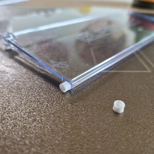 Könnte beinhalten: Ein durchsichtiges, rechteckiges Regalbauteil aus Kunststoff mit einer dünnen, transparenten Platte und einem Scharnier an einer Seite. Eine kleine, weiße zylindrische Kunststoffkappe steckt im Scharnier, eine zweite identische Kappe liegt daneben.