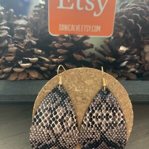 May include: A pair of teardrop-shaped dangle earrings with a brown and black snake skin pattern. The earrings are made of leather and have gold-colored hooks.