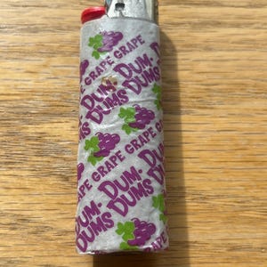 Puede incluir: Un encendedor plateado con un bot&oacute;n de encendido rojo. El encendedor est&aacute; envuelto en una etiqueta de papel con las palabras "DUM DUMS" y "GRAPE GRAPE" impresas, junto con im&aacute;genes de uvas moradas.