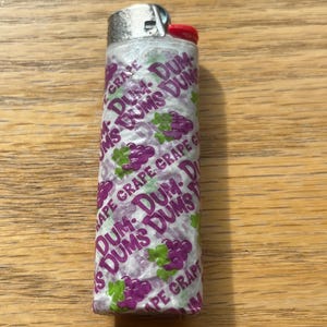 Puede incluir: Un encendedor desechable envuelto en un envoltorio de caramelo Dum Dums con sabor a uva. El envoltorio es blanco con texto morado e ilustraciones de uvas. El encendedor tiene una parte superior de metal plateado y un bot&oacute;n de encendido rojo.