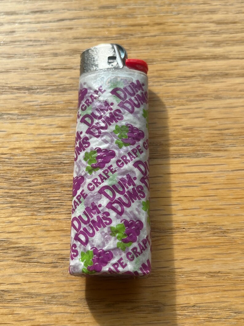 Puede incluir: Un encendedor desechable envuelto en un envoltorio de caramelo Dum Dums con sabor a uva. El envoltorio es blanco con texto morado e ilustraciones de uvas. El encendedor tiene una parte superior de metal plateado y un bot&oacute;n de encendido rojo.