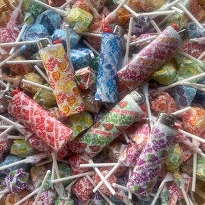 Puede incluir: Una cesta llena de coloridas piruletas Dum-Dums de varios sabores de frutas. Varios encendedores est&aacute;n esparcidos entre los caramelos. Las piruletas est&aacute;n envueltas en papel estampado con el logotipo de Dum-Dums e ilustraciones de frutas.