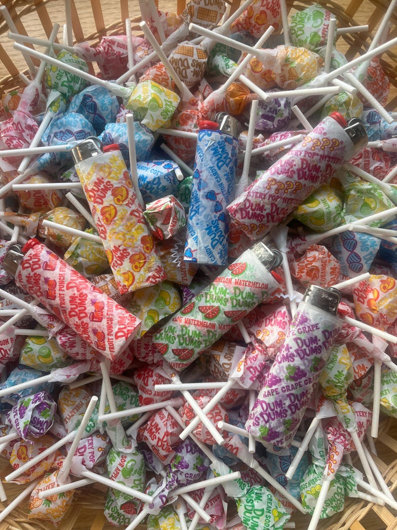 Puede incluir: Una cesta llena de coloridas piruletas Dum-Dums de varios sabores de frutas. Varios encendedores est&aacute;n esparcidos entre los caramelos. Las piruletas est&aacute;n envueltas en papel estampado con el logotipo de Dum-Dums e ilustraciones de frutas.