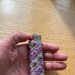 Puede incluir: Un encendedor plateado envuelto en papel blanco con las palabras "DUM-DUMS GRAPE" en morado y verde. El encendedor tiene un bot&oacute;n de encendido rojo.