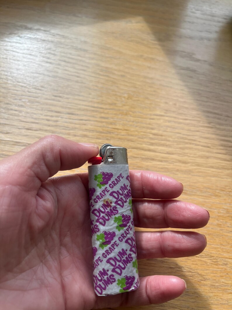 Puede incluir: Un encendedor plateado envuelto en papel blanco con las palabras "DUM-DUMS GRAPE" en morado y verde. El encendedor tiene un bot&oacute;n de encendido rojo.