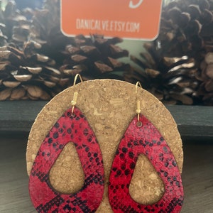 May include: A pair of red and black snake skin patterned dangle earrings with gold hooks. The earrings are shaped like teardrops with a cutout in the center.