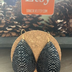 May include: A pair of black teardrop-shaped earrings with a snakeskin pattern. The earrings are made of leather and have gold-colored hooks.