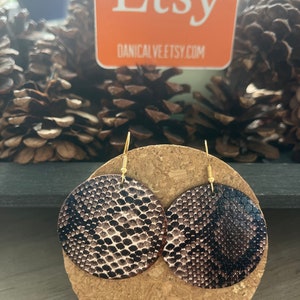 May include: A pair of round, brown and black snake skin patterned earrings with gold hooks. The earrings are displayed on a cork coaster.