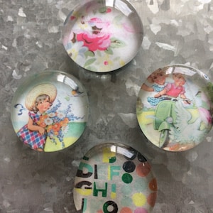 May include: Set of four glass paperweights with different vintage images. One features a floral pattern with pink roses, another shows a girl with a bouquet of flowers, a third depicts two children riding a scooter, and the last one has a colorful abstract design with letters and dots.