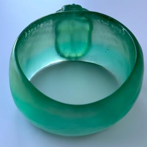May include: A teal-coloured bangle bracelet. The bracelet is translucent and has a rounded shape with a smooth, glossy finish. The words "Come into" are etched on the inside. There is a small, raised design on the top of the bracelet.
