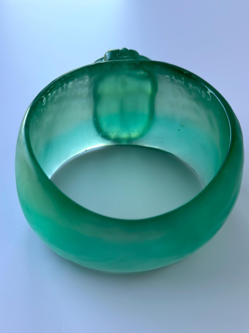 May include: A teal-coloured bangle bracelet. The bracelet is translucent and has a rounded shape with a smooth, glossy finish. The words "Come into" are etched on the inside. There is a small, raised design on the top of the bracelet.