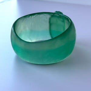 May include: A teal-coloured glass bracelet. The bracelet is a thick, rounded band with a smooth, polished surface. The colour transitions from a deeper teal to a lighter, almost seafoam green hue. The bracelet is a solid, circular shape.