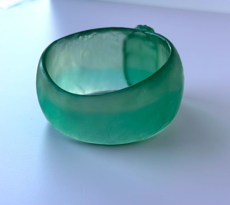 May include: A teal-coloured glass bracelet. The bracelet is a thick, rounded band with a smooth, polished surface. The colour transitions from a deeper teal to a lighter, almost seafoam green hue. The bracelet is a solid, circular shape.