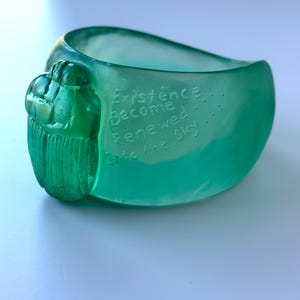 May include: A teal-coloured ring with a scarab beetle design on the side. The ring has the words "Existence Become Renewed Into the Sky" etched into the surface. The ring is made of a smooth, glossy material.