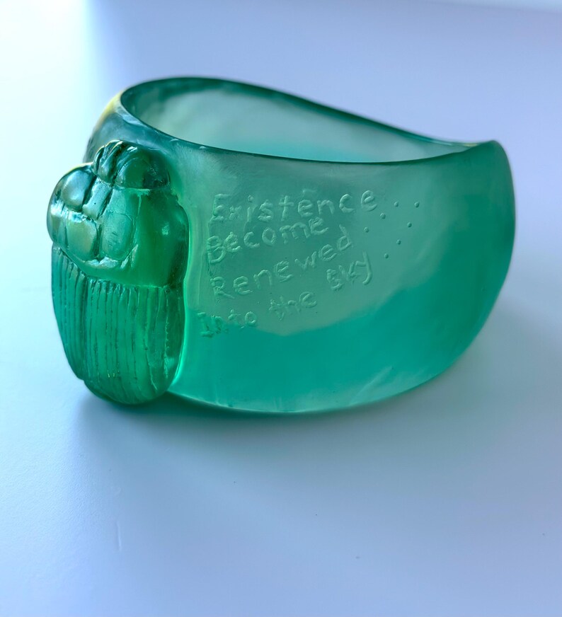 May include: A teal-coloured ring with a scarab beetle design on the side. The ring has the words "Existence Become Renewed Into the Sky" etched into the surface. The ring is made of a smooth, glossy material.