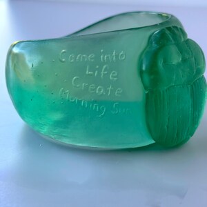 May include: A teal-coloured glass bowl with the words "Come into Life Create Morning Sun" etched into the side. The bowl has a rounded shape with a textured, carved design on one side.