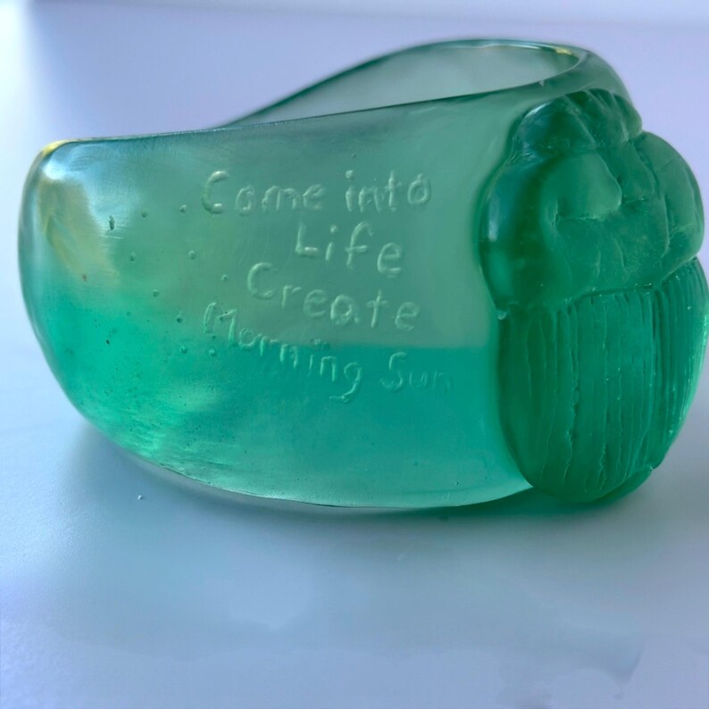 May include: A teal-coloured glass bowl with the words "Come into Life Create Morning Sun" etched into the side. The bowl has a rounded shape with a textured, carved design on one side.