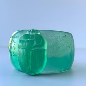 May include: A translucent green glass bowl with a raised scarab beetle design. The bowl has etched text that reads "Into life create shining sun" and "Existence Become Renewed Into the sky".