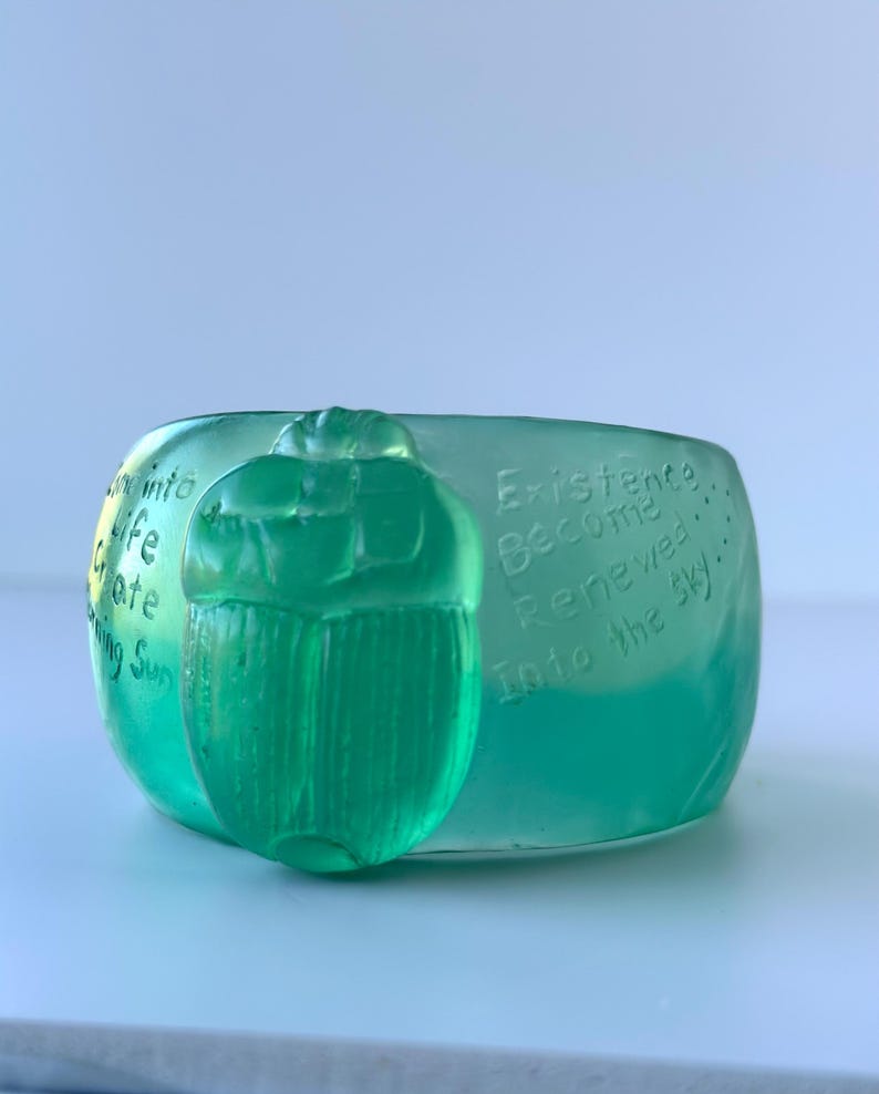 May include: A translucent green glass bowl with a raised scarab beetle design. The bowl has etched text that reads "Into life create shining sun" and "Existence Become Renewed Into the sky".