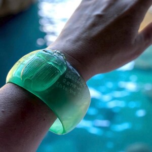 May include: A vibrant green cuff bracelet featuring a carved scarab beetle. The bracelet has engraved text that reads "Become Renew Into the". The bracelet is worn on a wrist, with a blurred blue background.