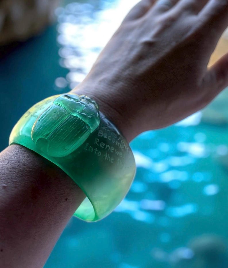 May include: A vibrant green cuff bracelet featuring a carved scarab beetle. The bracelet has engraved text that reads "Become Renew Into the". The bracelet is worn on a wrist, with a blurred blue background.