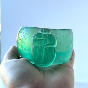 May include: A translucent green glass bowl with a scarab beetle design. The bowl has etched text that reads "Come into Life, Create, Morning Sun" and "Existence Become Renewed Into the Sky".