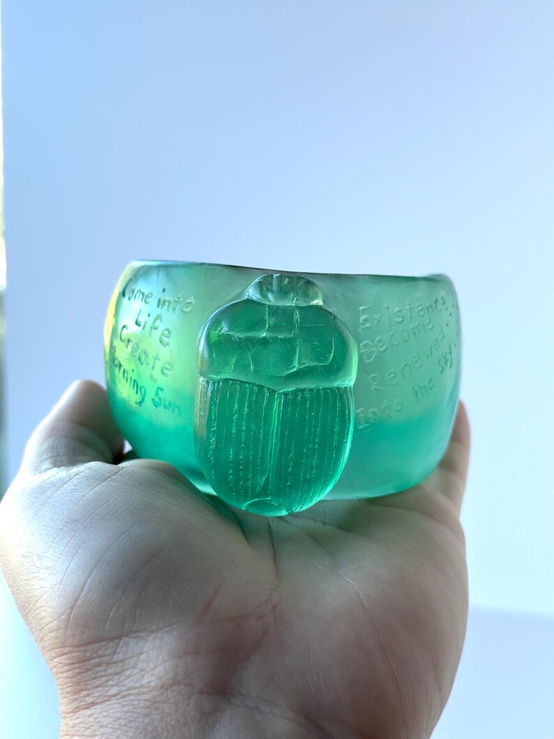 May include: A translucent green glass bowl with a scarab beetle design. The bowl has etched text that reads "Come into Life, Create, Morning Sun" and "Existence Become Renewed Into the Sky".