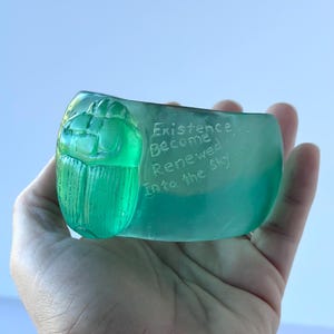 May include: A hand holds a translucent, teal-coloured glass sculpture. The sculpture features a detailed scarab beetle design on one end and the words "Existence Become Renewed Into the Sky" etched on the side.