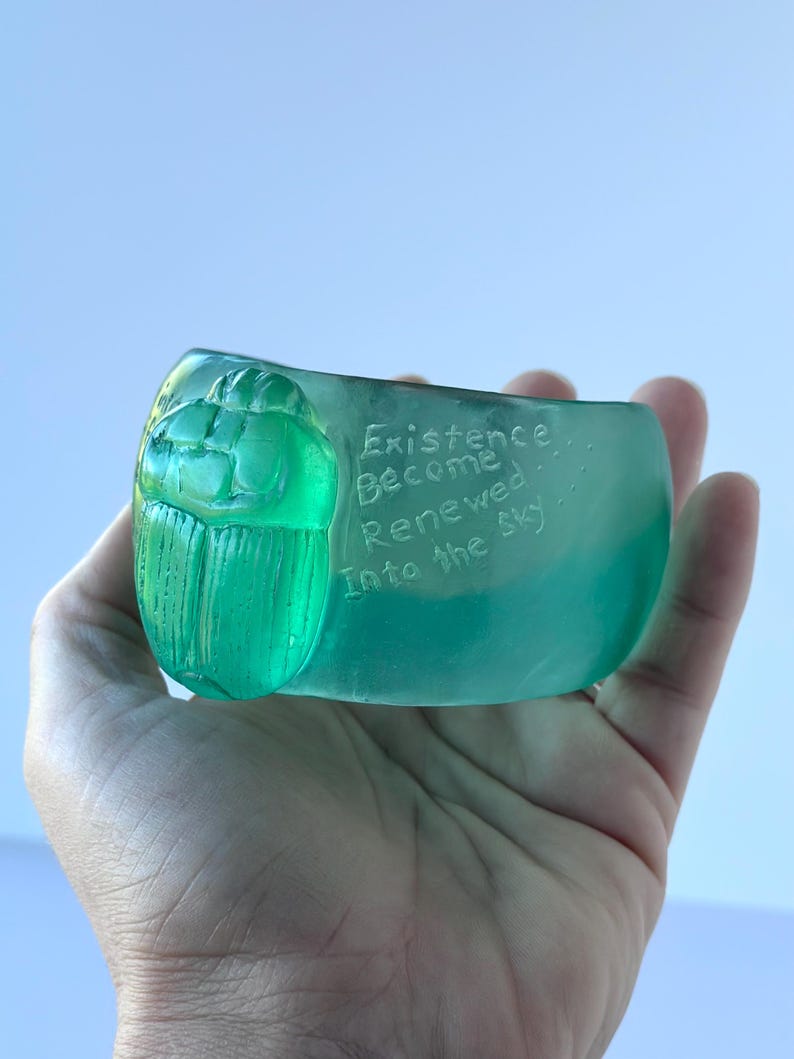 May include: A hand holds a translucent, teal-coloured glass sculpture. The sculpture features a detailed scarab beetle design on one end and the words "Existence Become Renewed Into the Sky" etched on the side.