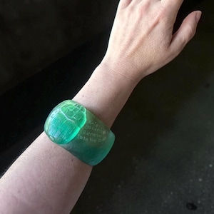 May include: A translucent green cuff bracelet with engraved text. The bracelet is a vibrant shade of green and appears to be made of a solid material. The text on the bracelet reads "Existing Becomes Renewed Into the Sky."