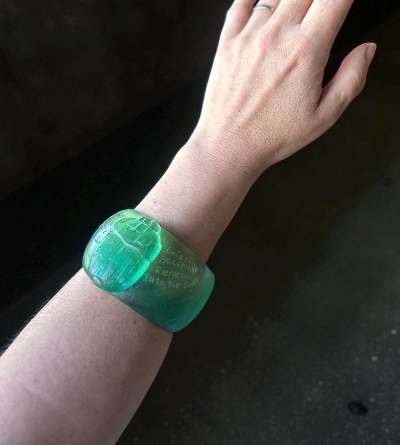 May include: A translucent green cuff bracelet with engraved text. The bracelet is a vibrant shade of green and appears to be made of a solid material. The text on the bracelet reads "Existing Becomes Renewed Into the Sky."