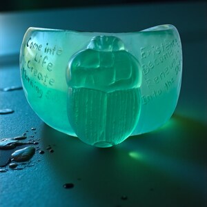 May include: A teal-coloured bowl with a carved scarab beetle design. The bowl has etched text that reads "Come into Life Create Morning &" on one side and "Existence Become Renewed Into the Sky" on the other.