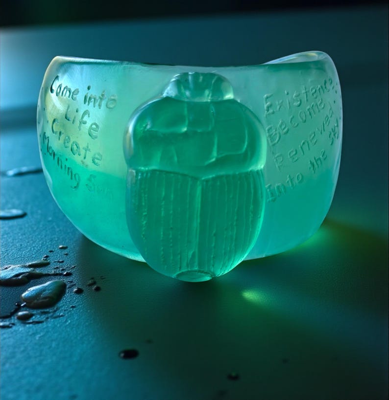 May include: A teal-coloured bowl with a carved scarab beetle design. The bowl has etched text that reads "Come into Life Create Morning &" on one side and "Existence Become Renewed Into the Sky" on the other.