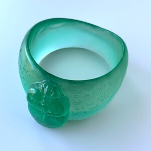 May include: A translucent teal bracelet with a rounded shape. The bracelet has a raised, carved design and etched text. The text reads "Existence Becomes Renewed Into the Sky".