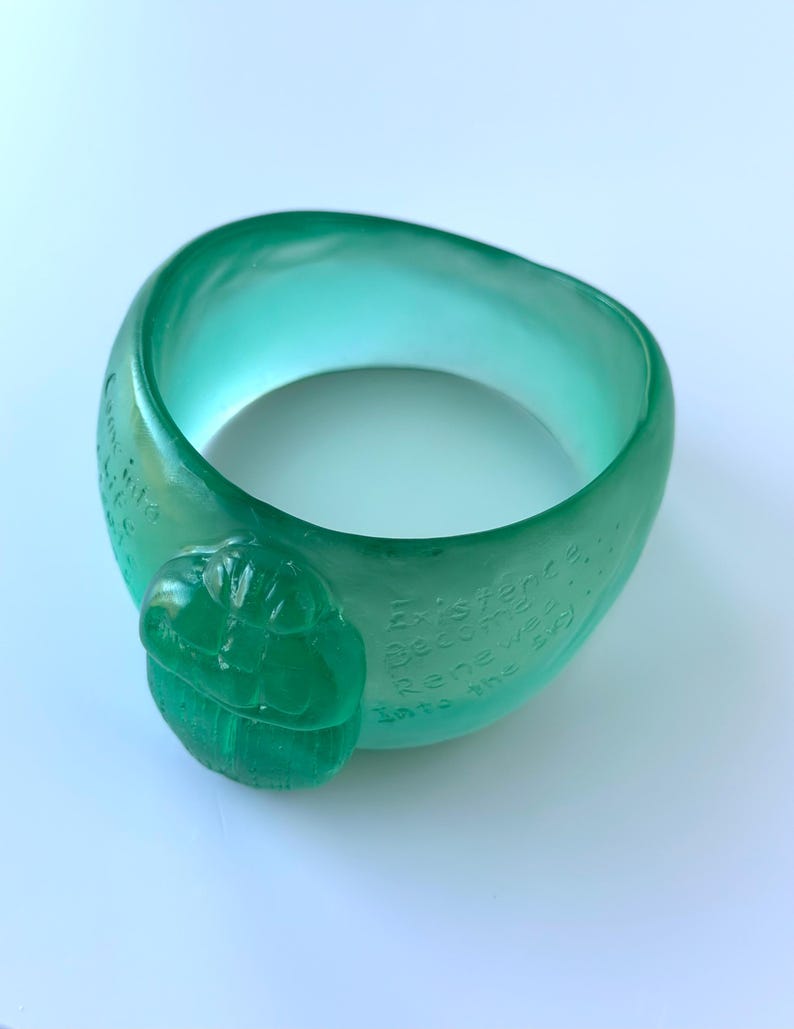 May include: A translucent teal bracelet with a rounded shape. The bracelet has a raised, carved design and etched text. The text reads "Existence Becomes Renewed Into the Sky".