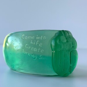 May include: A translucent green decorative object with the words "Come into Life Create Morning Sun" etched into the surface. The object has a rounded shape with a detailed, carved beetle design on one end.