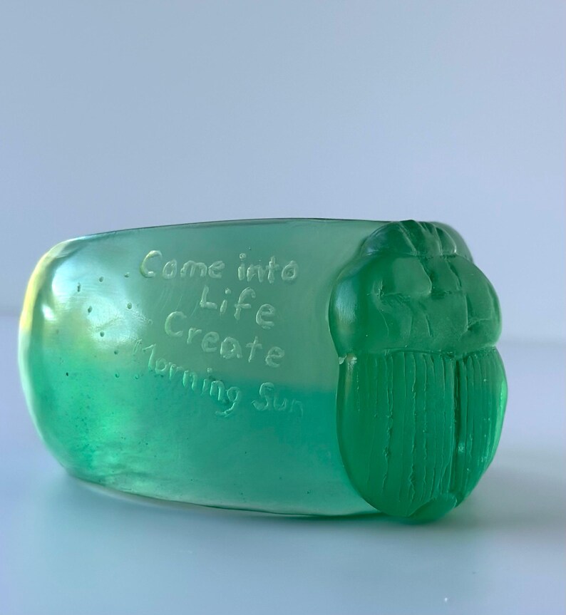May include: A translucent green decorative object with the words "Come into Life Create Morning Sun" etched into the surface. The object has a rounded shape with a detailed, carved beetle design on one end.