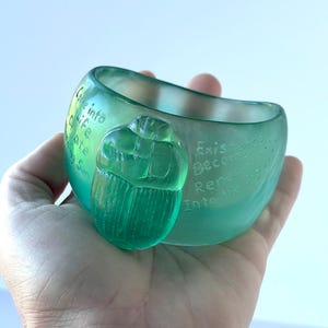 May include: A translucent teal glass bowl with a carved scarab beetle. The bowl has etched text reading "Come into Life, Create, Rising Sun" and "Existence Become Renew Into the Sky."