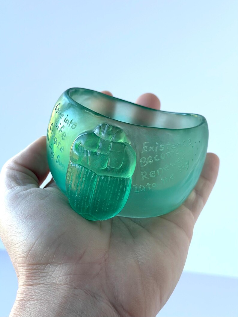 May include: A translucent teal glass bowl with a carved scarab beetle. The bowl has etched text reading "Come into Life, Create, Rising Sun" and "Existence Become Renew Into the Sky."