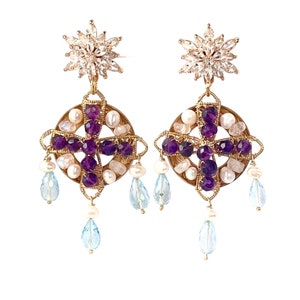 May include: Gold tone earrings with a purple amethyst and white pearl design. The earrings feature a starburst charm at the top and dangle with blue teardrop stones.