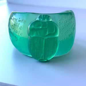May include: A teal-coloured bowl with a scarab beetle design. The bowl has etched text that reads "Come into Life Create Morning Sun" and "Existence Become Renewed Into the Sky."