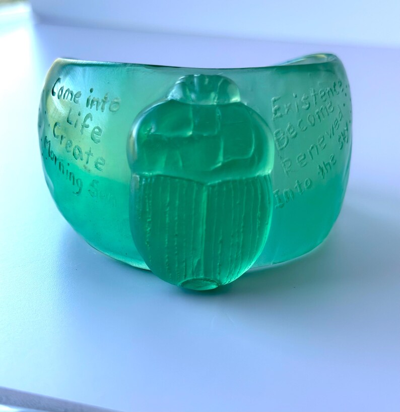 May include: A teal-coloured bowl with a scarab beetle design. The bowl has etched text that reads "Come into Life Create Morning Sun" and "Existence Become Renewed Into the Sky."