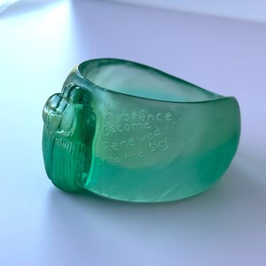 May include: A teal-coloured glass bracelet with a textured, organic design. The bracelet features the etched words "Existence Become Renewed Into the Sky." The bracelet has a rounded shape and a decorative, textured element on one side.