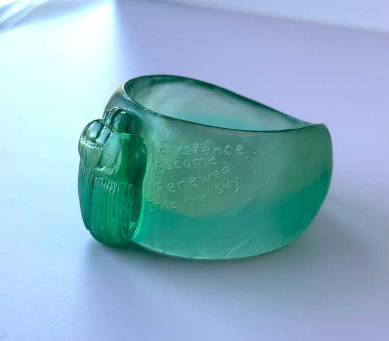 May include: A teal-coloured glass bracelet with a textured, organic design. The bracelet features the etched words "Existence Become Renewed Into the Sky." The bracelet has a rounded shape and a decorative, textured element on one side.