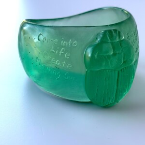 May include: A translucent green glass bowl with the words "Come into Life Create Morning Sun" etched on the side. A green scarab beetle is on the side of the bowl.