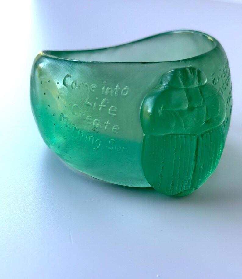 May include: A translucent green glass bowl with the words "Come into Life Create Morning Sun" etched on the side. A green scarab beetle is on the side of the bowl.