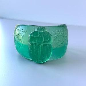 May include: A translucent green bowl with a scarab beetle design. The bowl has etched text that reads "Come into Life, Create, Morning Sun" and "Existence Become Renewed Into the 3D."