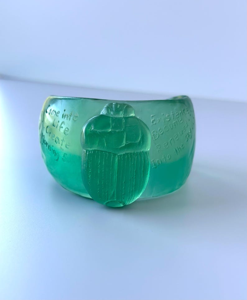 May include: A translucent green bowl with a scarab beetle design. The bowl has etched text that reads "Come into Life, Create, Morning Sun" and "Existence Become Renewed Into the 3D."